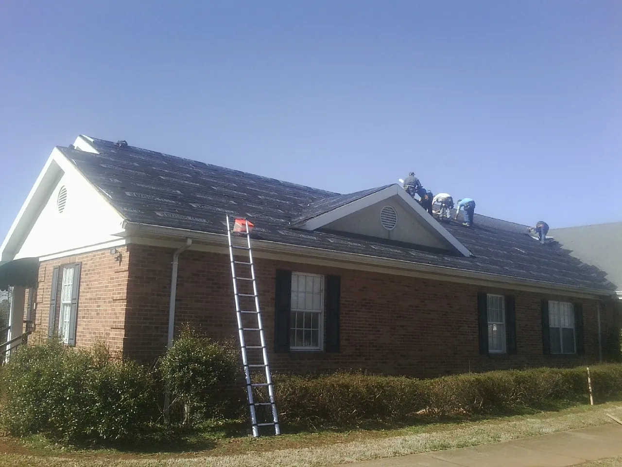 Crew tearing shingles off a brick ranch roof with a ladder set — Metal Roof Repair work by our crew in Lacon