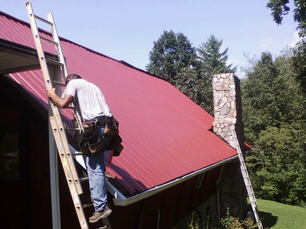 Roofer on a ladder beside a red metal roof and stone chimney — Soffit & Fascia Repair by Roofing Brothers in Lacon